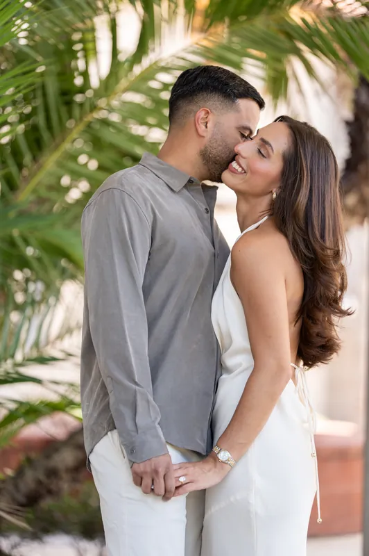 Nestled among palm branches, one partner kisses the other's cheek while they hold hands, sunlight filtering through green leaves.