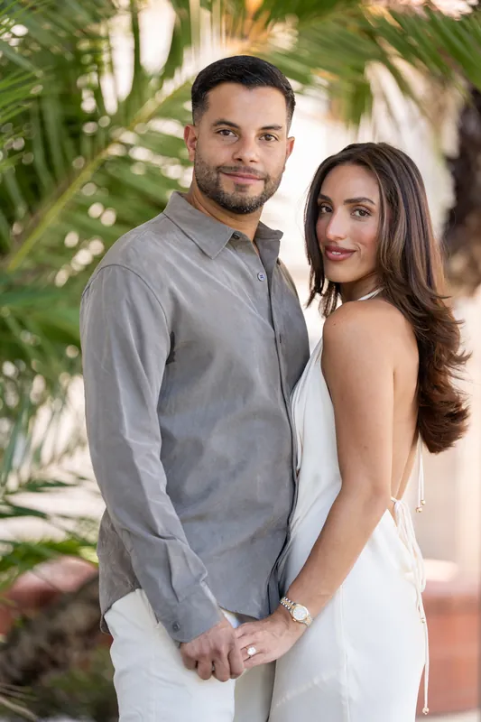 The couple holds hands and faces the camera with relaxed smiles, palm fronds softly blurred in the background of the museum courtyard.