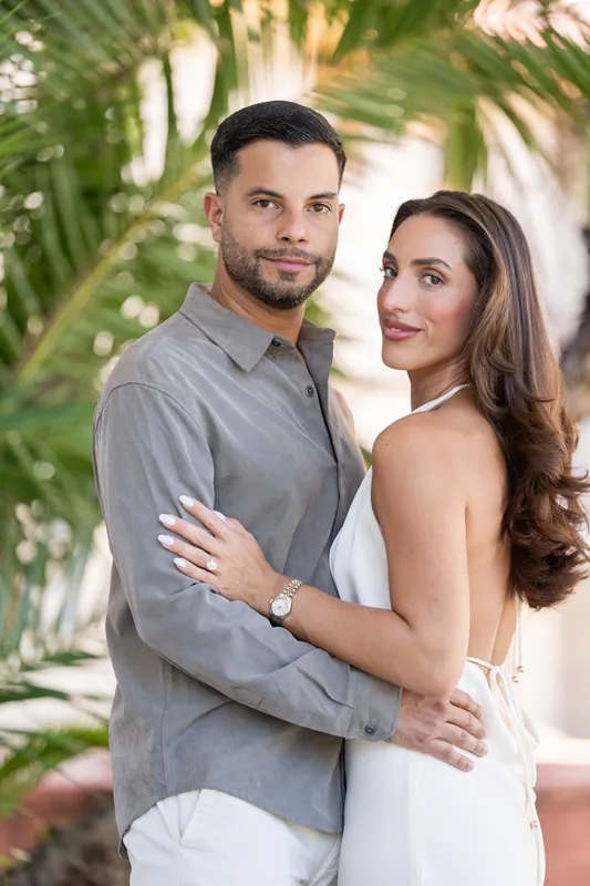 Two people embrace under tropical palms at the Lightner Museum Courtyard, one in gray and one in white, both smiling warmly.