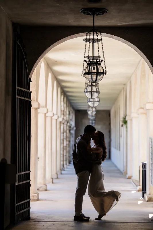 Two figures embrace in a long white corridor lined with columns and a hanging lantern at the Lightner Museum Courtyard.