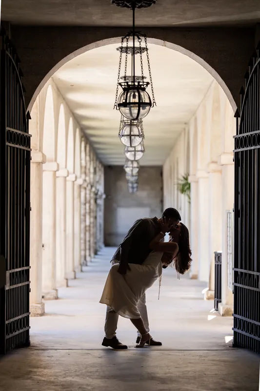 Someone in dark clothing dips their partner in the colonnade, pale columns stretching behind them under an ornate ceiling fixture.