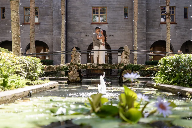 A couple stands on a balcony overlooking the museum's central courtyard fountain, framed by palm trees and flowering plants below.