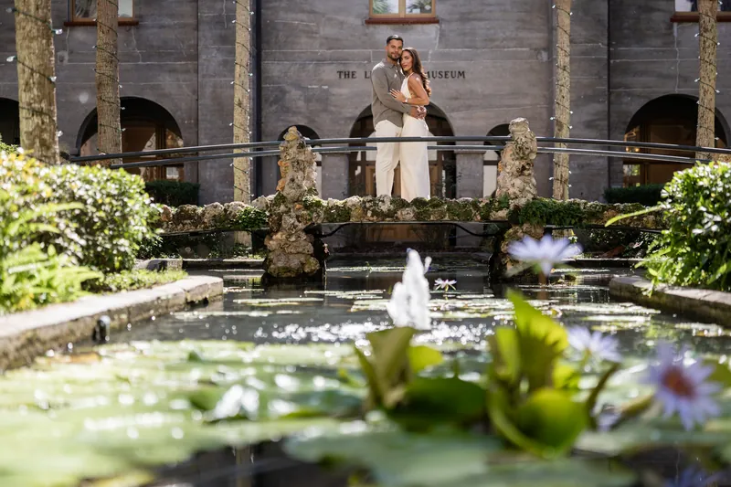 Two people stand together on a balcony railing overlooking a reflective pool with water lilies and blooming flowers at the Lightner Museum.