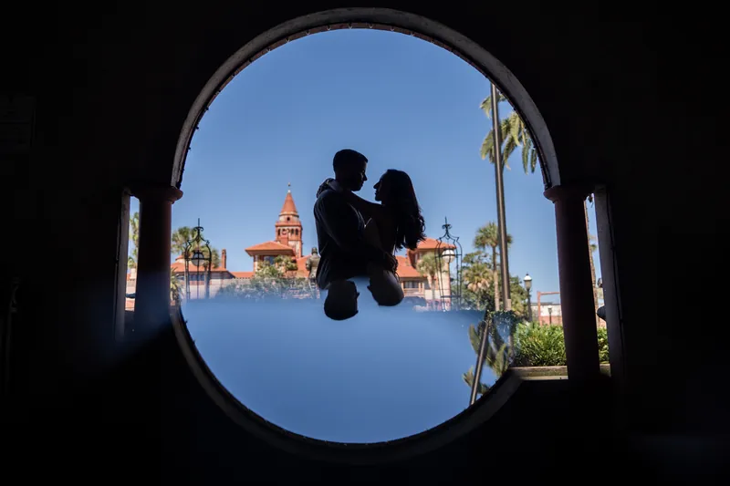 Two people embrace in silhouette within a circular stone frame at the Lightner Museum, with a cathedral dome visible beyond.