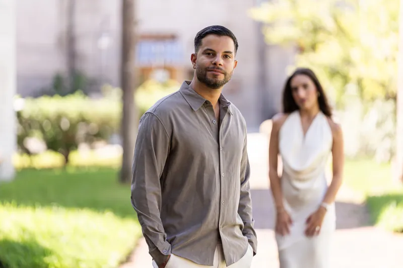 A person in a gray shirt walks forward smiling while another in a white sleeveless top follows behind in a sunny courtyard setting.