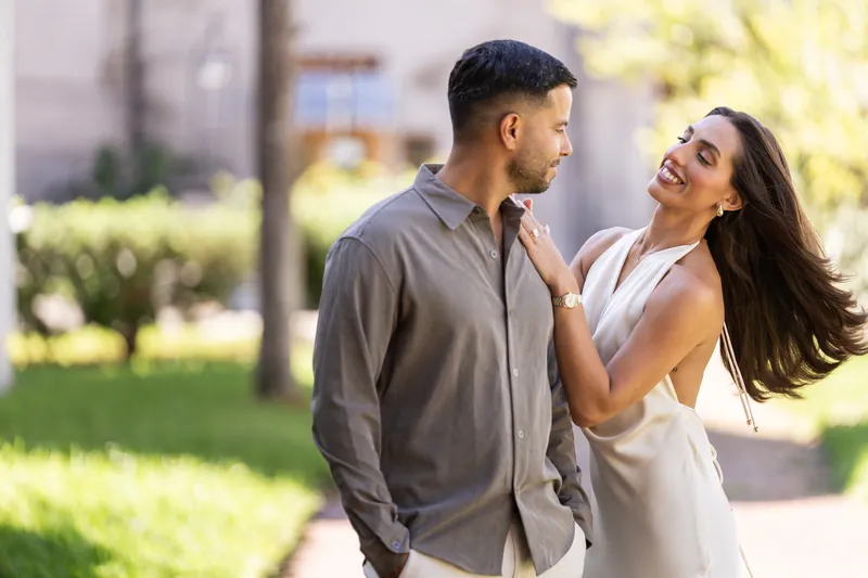 Two people stand close together in dappled sunlight, one reaching toward the other's shoulder with a joyful expression at the museum courtyard.