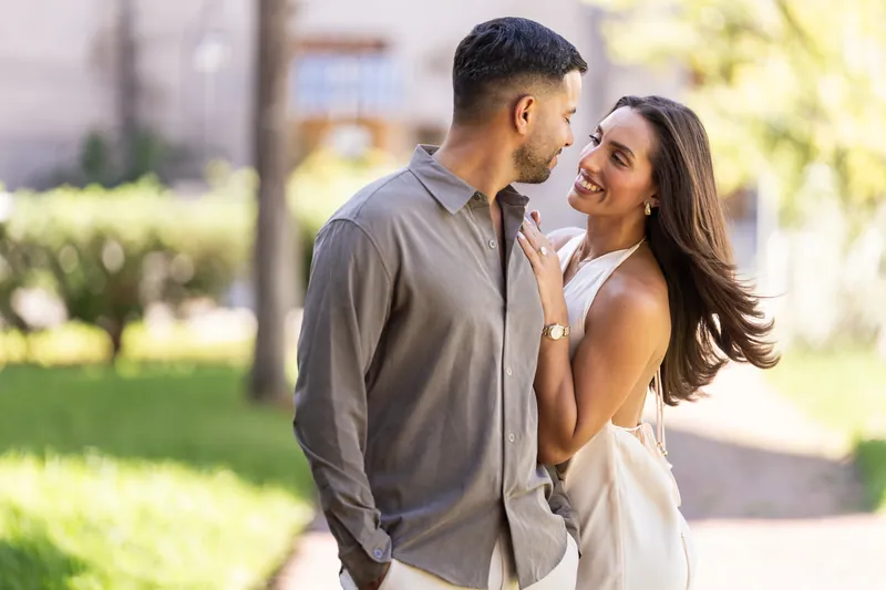 A couple holds each other while laughing in the bright Palm Row gardens, with lush greenery softly visible in the background.