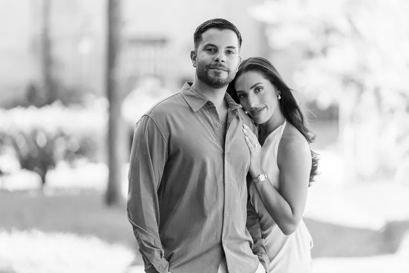 Two people pose together in black and white, leaning into each other with relaxed warmth against an architectural backdrop.