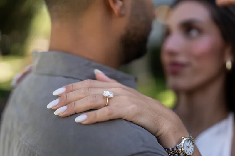A diamond engagement ring sparkles on someone's hand as they rest against their partner's shoulder in soft afternoon light.