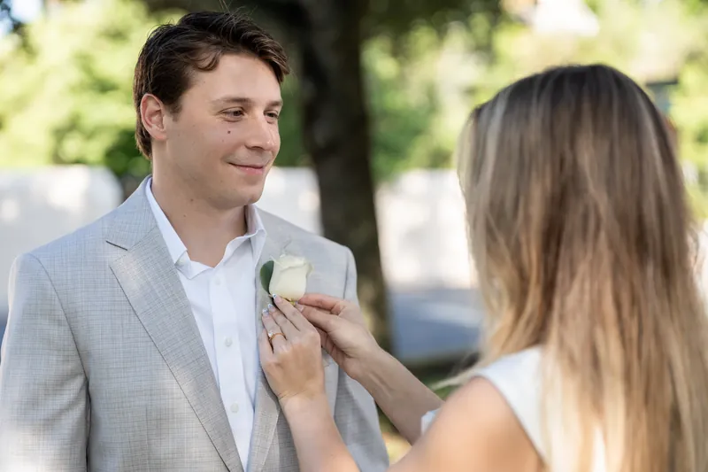 Devin smiles while Blair fastens a white flower to his light gray jacket lapel.
