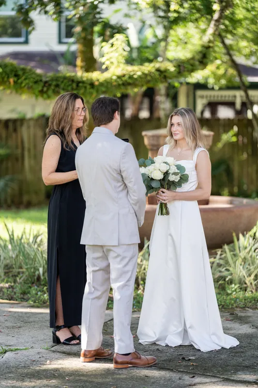 An officiant in black speaks with Devin and Blair, who holds a white flower bouquet, during their ceremony.