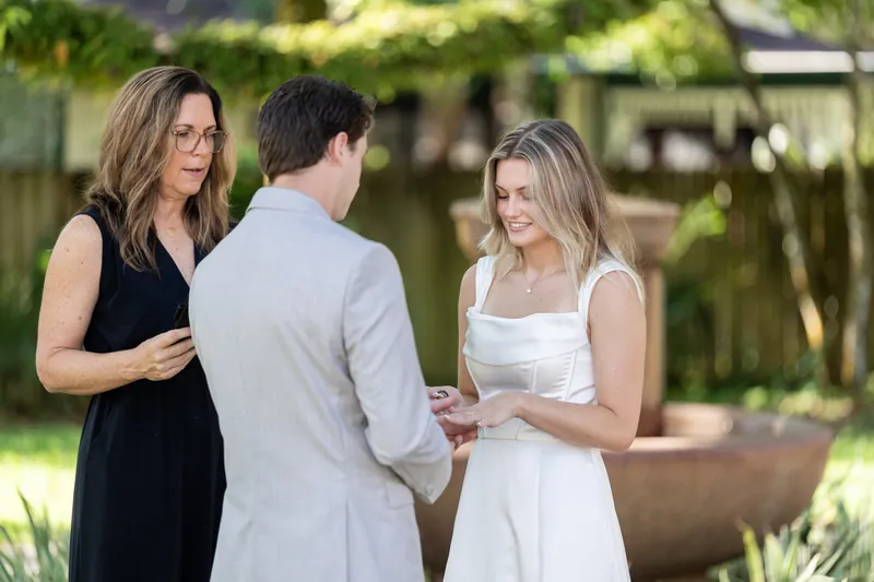 An officiant in black stands between Devin and Blair as they hold hands during their ceremony under shade.