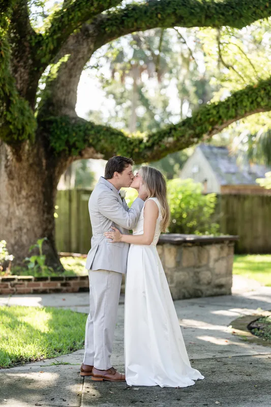 Devin and Blair kiss beneath an ancient moss-draped oak tree, with a stone structure visible behind them at St. Francis Park.