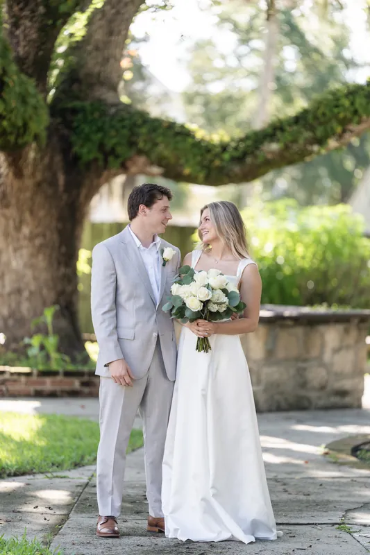 Blair holds a white flower bouquet while standing beside Devin in his gray suit under the sprawling oak at St. Francis Park.