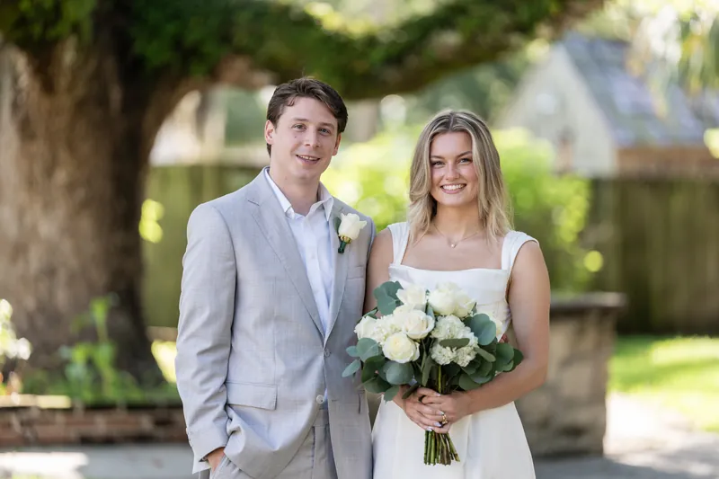 Devin and Blair walk arm-in-arm through sunlit grounds, Blair holding a white bouquet.