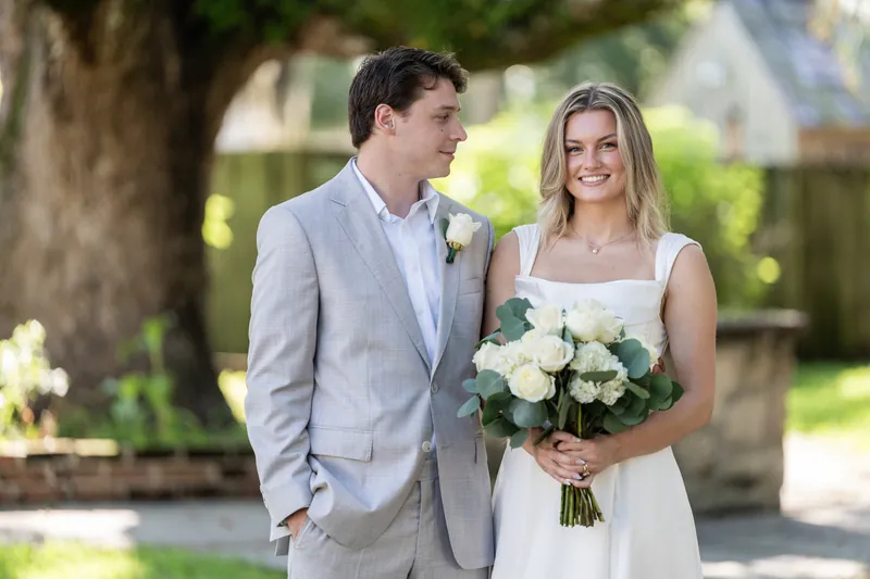Devin glances at Blair as they walk together holding hands, both smiling, with her white bouquet visible at St. Francis Park.