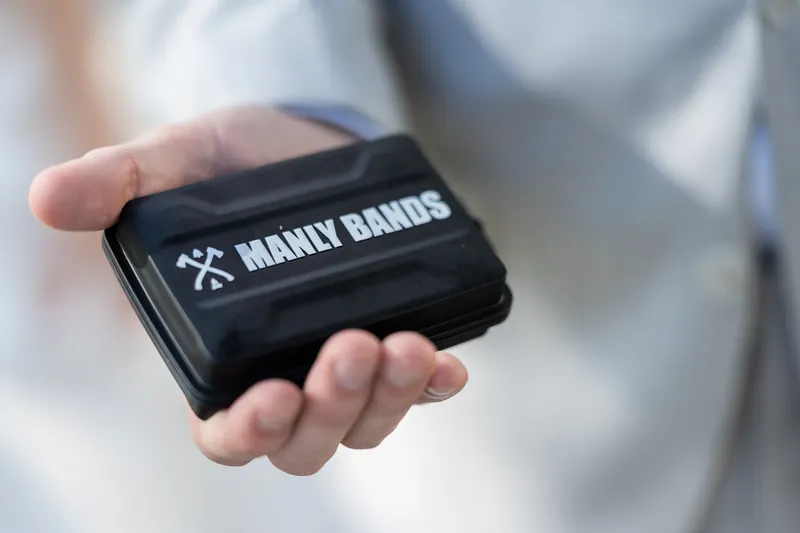Devin's hand holds a black wallet embossed with "MANLY BANDS" while wearing a white dress shirt.