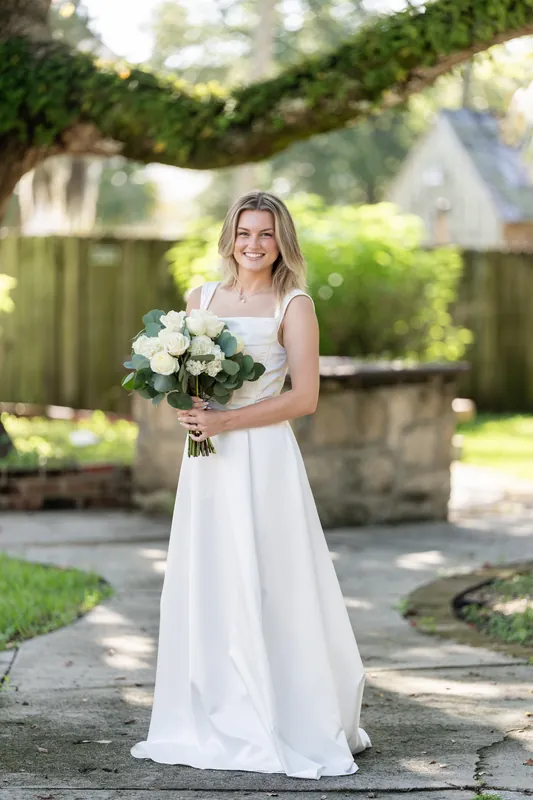 Blair stands alone on a stone pathway at St. Francis Park, holding a white rose bouquet and wearing a flowing white gown.