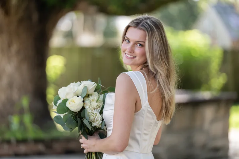 Blair smiles over her shoulder while holding a white rose bouquet in her sleeveless dress.