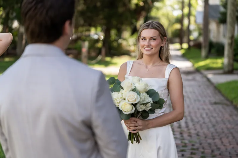 Blair faces Devin on a palm-lined street, holding her bouquet while Devin stands in profile wearing a light gray suit.