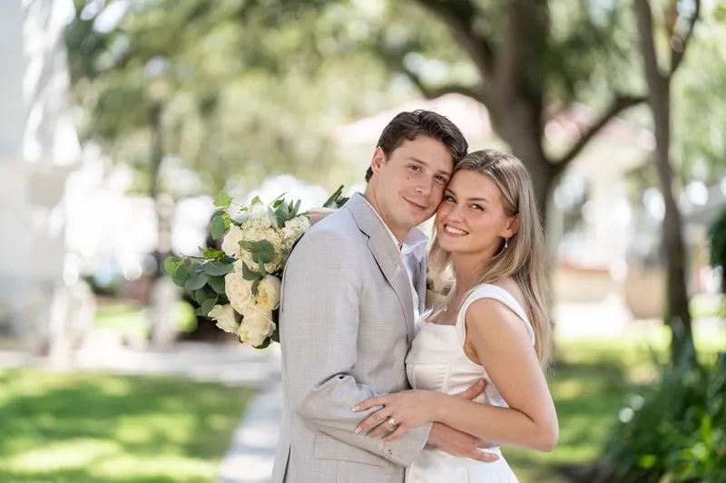 Blair leans into Devin's embrace while holding a white bouquet in the dappled shade of the plaza.