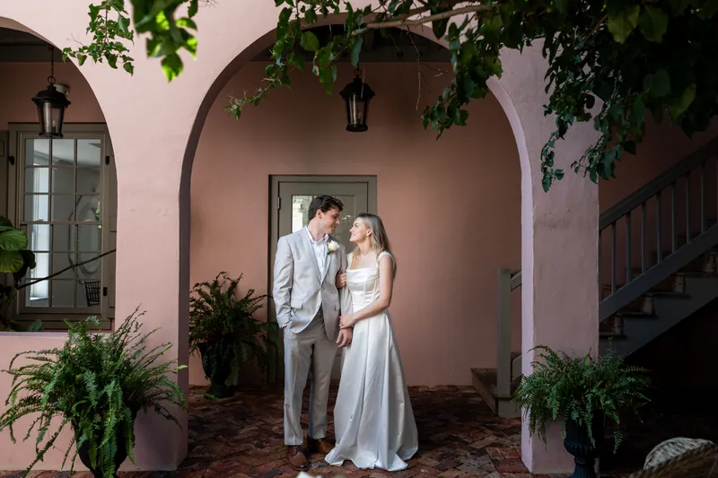 Blair and Devin stand together beneath a pink arched alcove decorated with climbing vines and potted plants.