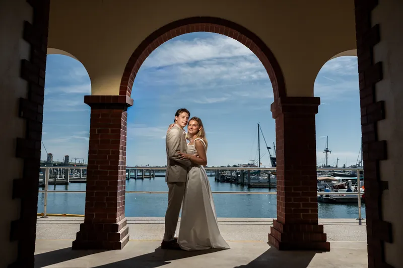 Blair and Devin embrace under a brick archway with sailboats and the St. Augustine Bayfront framed behind them.