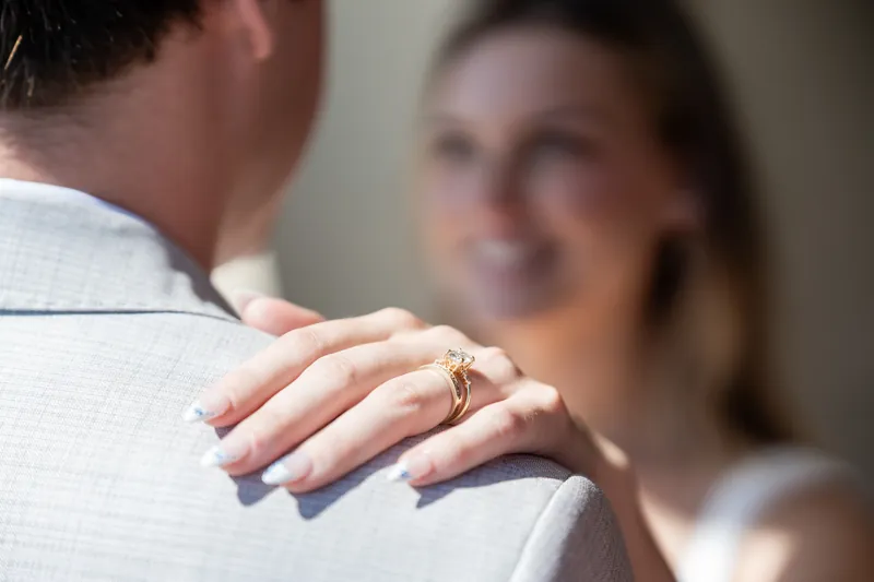 Devin's hand displays a wedding band while Blair smiles softly in the background during an intimate indoor moment.