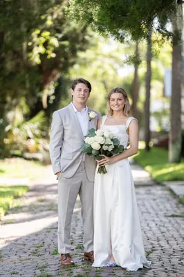 Blair and Devin walk arm-in-arm down a tree-lined brick path at Palm Row, with white flowers in hand.