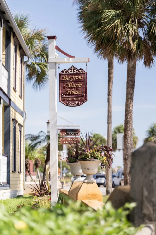 Brown wooden signage for Bayfront Marin House hangs from a white building, with palm trees and blue sky visible beyond.