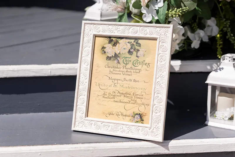 A framed cream-colored wedding program decorated with floral illustrations rests on a table surrounded by white flowers.