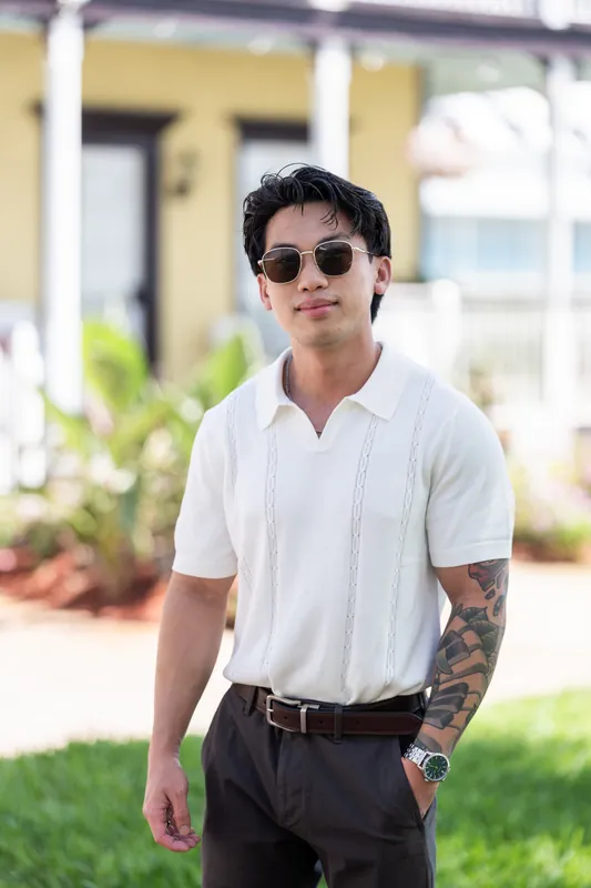 Chris stands on the lawn wearing sunglasses, a white short-sleeve shirt, and dark trousers, with a visible arm tattoo.