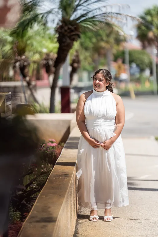 Blossom walks along a shaded sidewalk in a sleeveless white knee-length dress with hands clasped in front.
