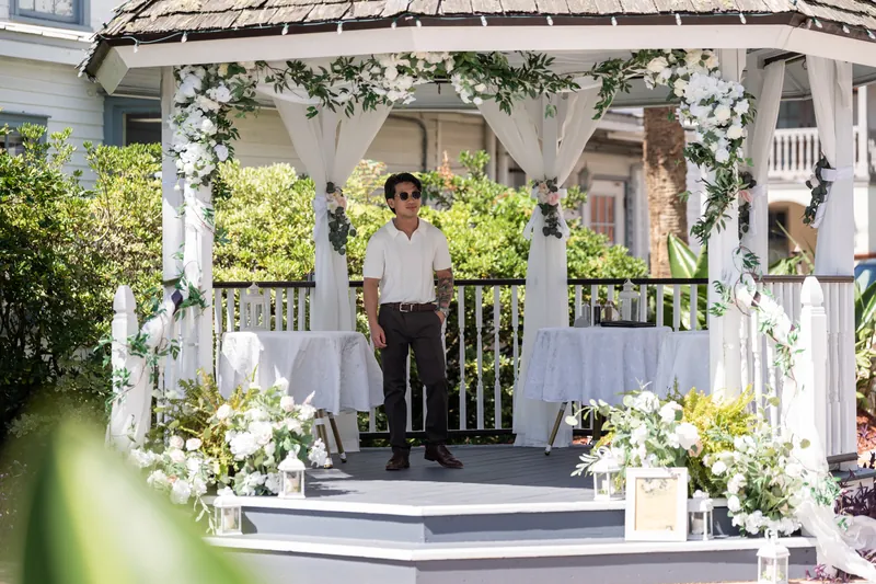 Chris stands inside the white gazebo under draped greenery and white florals, beside Blossom in her white dress.