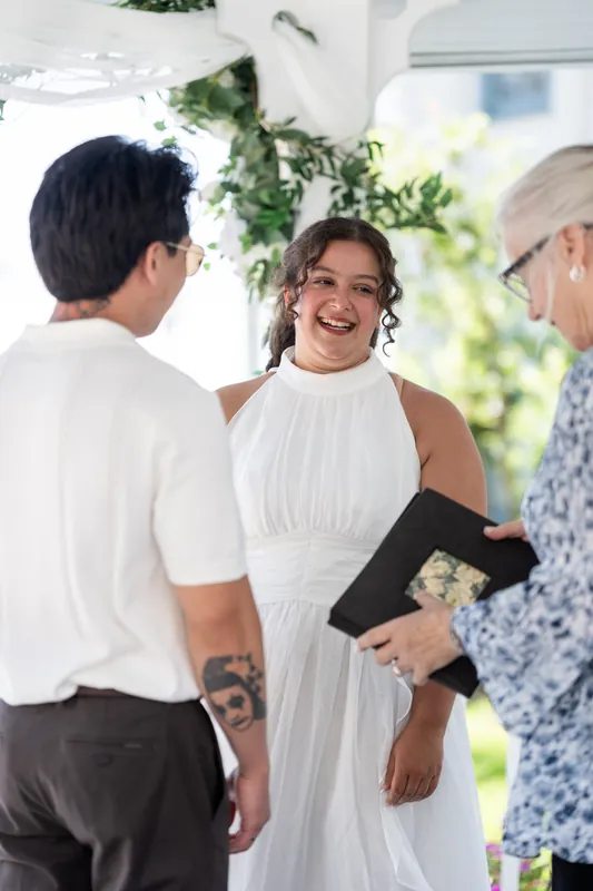Blossom smiles warmly while standing between Chris and an officiant holding a black book during their ceremony.