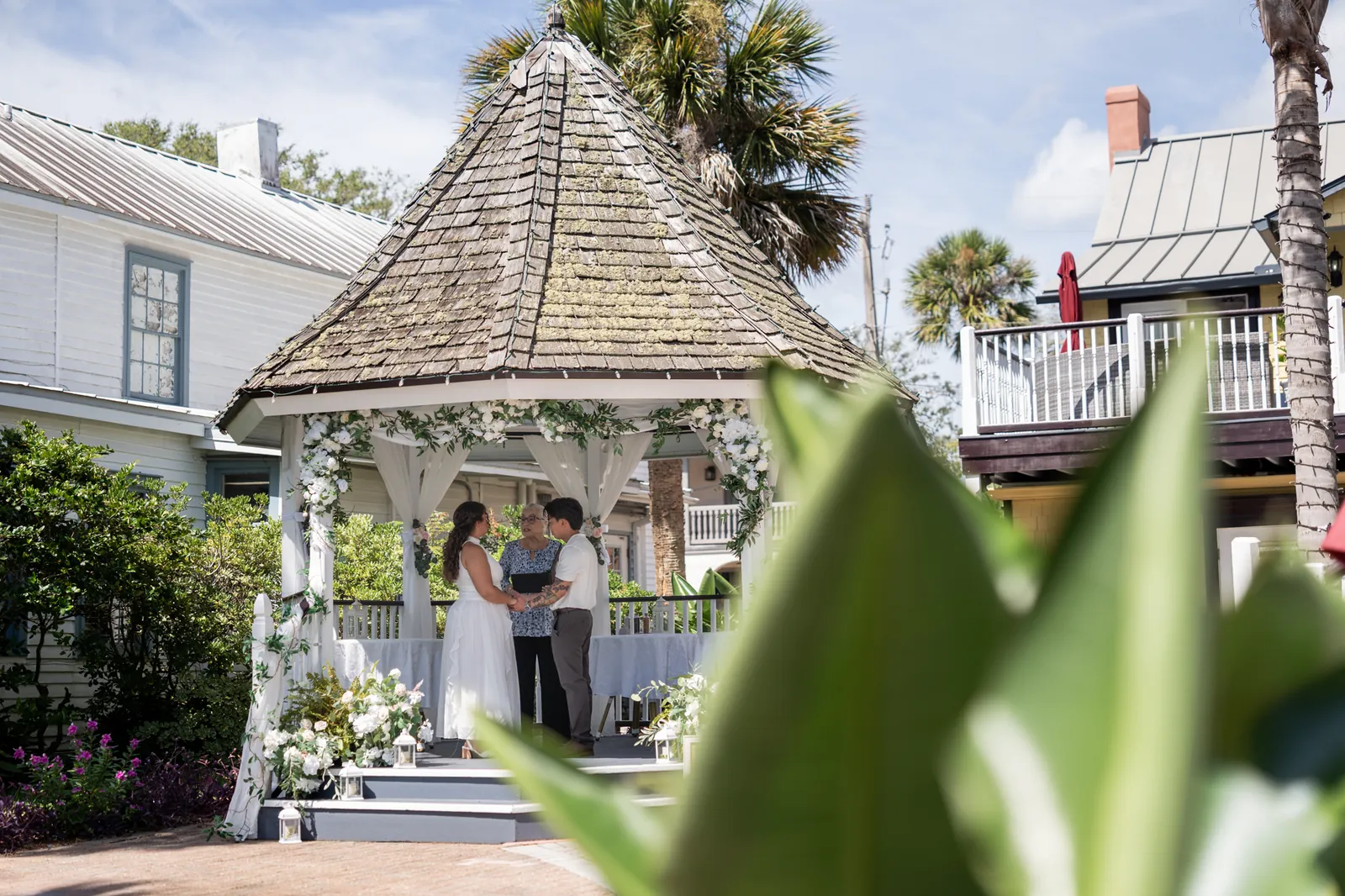 The couple stands under a peaked-roof gazebo with ivy garland while guests look on across the manicured grounds.