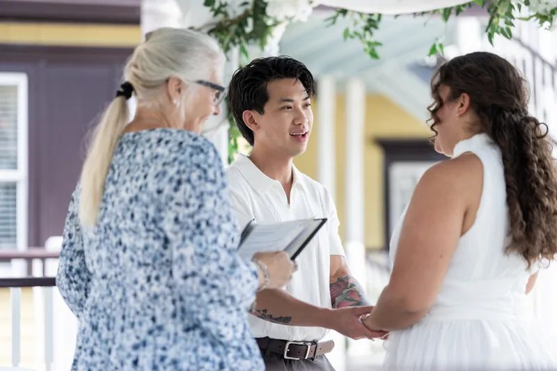 The officiant presents Blossom and Chris to their gathered guests with hands clasped together on the covered porch.