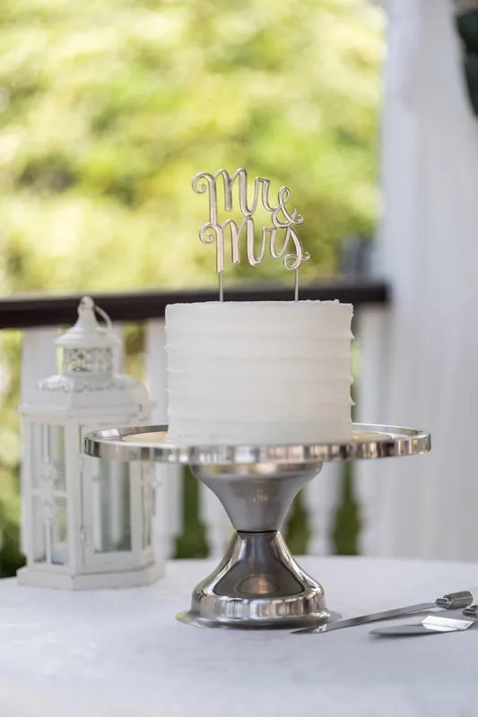 A tiered white cake topped with a "Mr. Mrs." gold topper sits on a pedestal table with champagne glasses nearby.