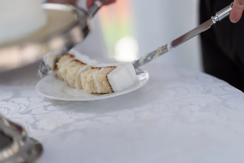 Two cake servers lift a slice of white cake with yellow filling onto a white plate during the reception.