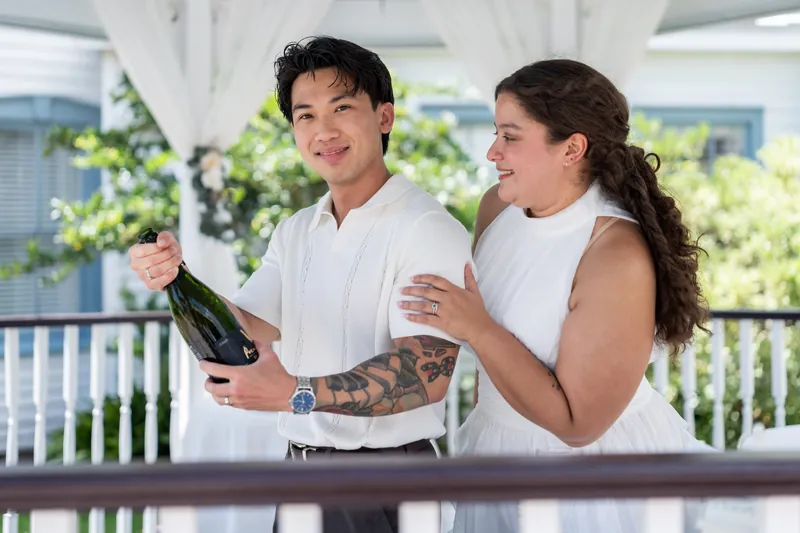 Chris and Blossom toast with champagne and cider on the covered veranda, both dressed in white and grinning at each other.