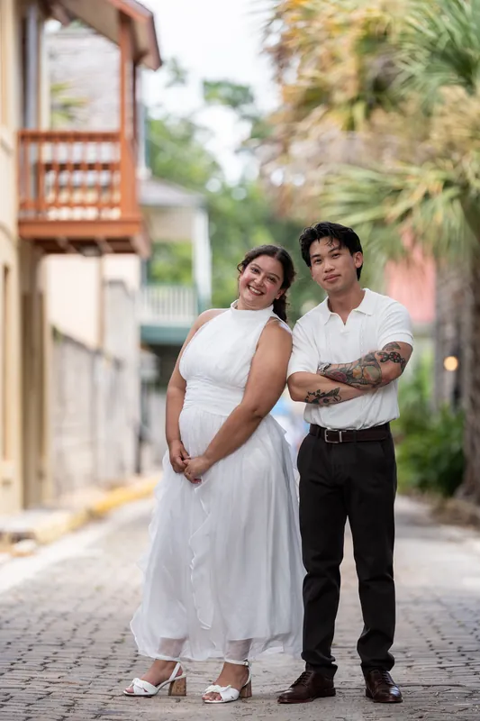 Blossom wears a flowing white dress while Chris stands beside her in a white shirt and black pants on a cobblestone street