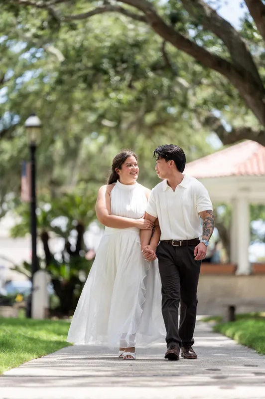 The couple strolls beneath spreading oak branches on a shaded plaza in downtown St. Augustine, holding hands as they walk.