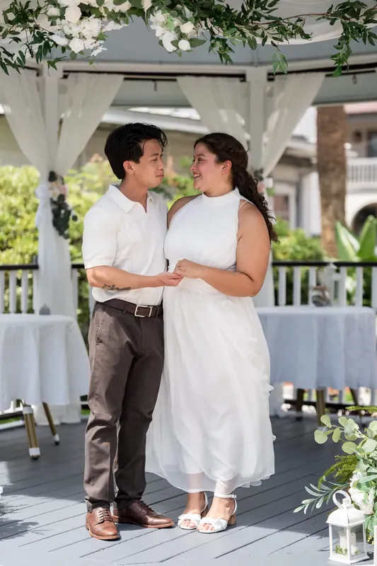 The couple leans close under draped white fabric on the deck, framed by flowering greenery.