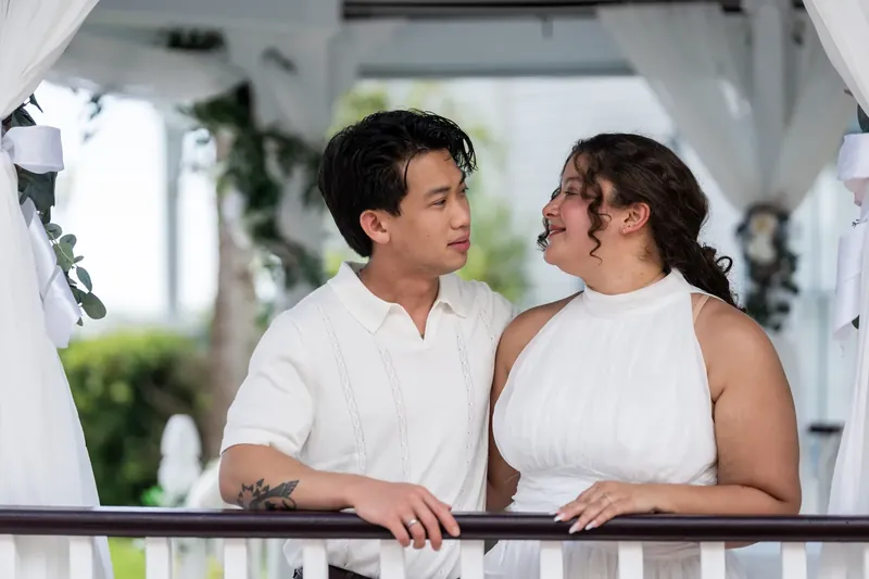Two people rest their arms on the white railing, gazing at each other beneath the pergola.