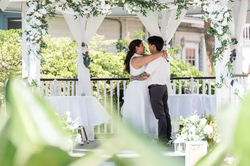 Two people embrace under a white tulle archway decorated with white blooms at the ceremony.