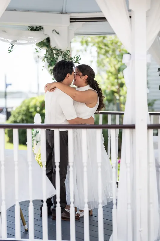 Blossom and Chris embrace on a white-railed balcony at Bayfront Marin House, with green water and trees visible behind them.