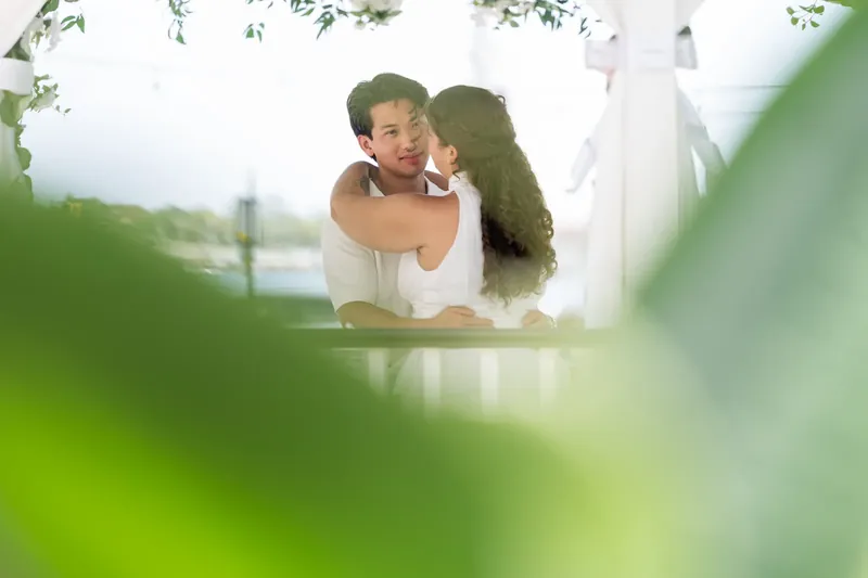 Framed through soft green foliage, one partner kisses the other's cheek while they stand together at the venue.
