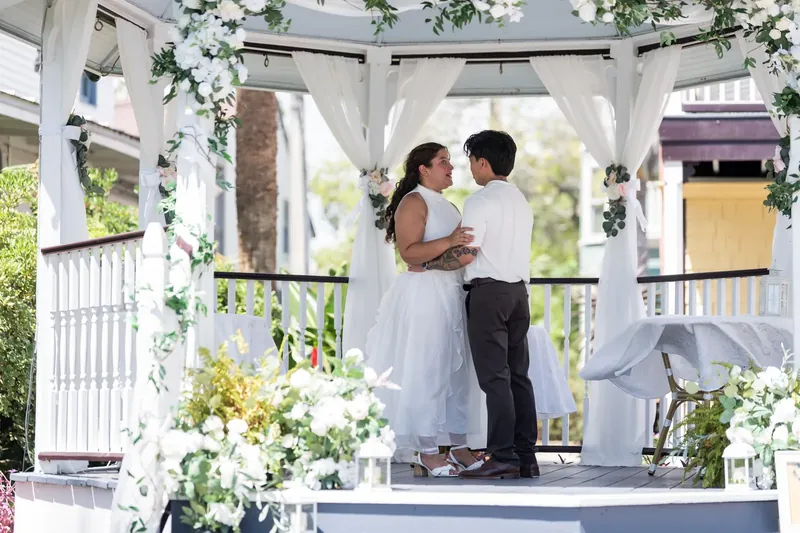 Two people stand face-to-face beneath a flowering white pergola, exchanging vows at the venue.