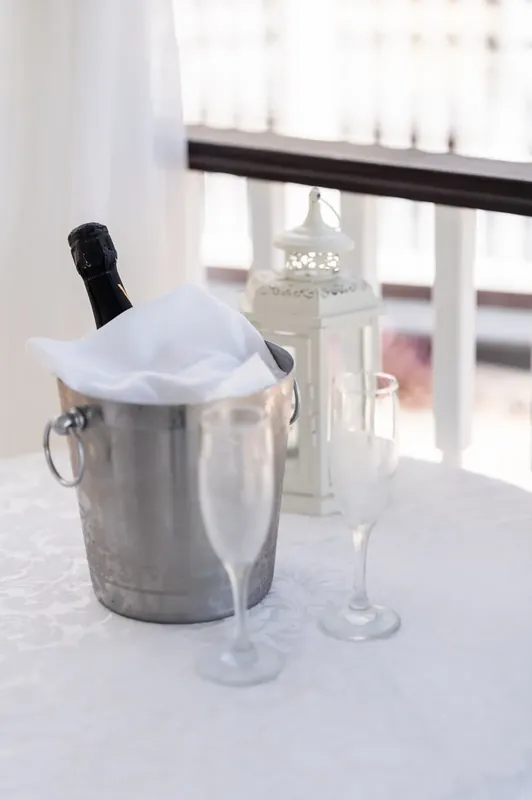 Champagne chills in a metal ice bucket next to a flute glass on a white-draped table with bay views.