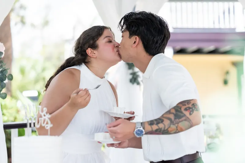Blossom and Chris lean in for a kiss while holding cake and champagne flutes at their reception.
